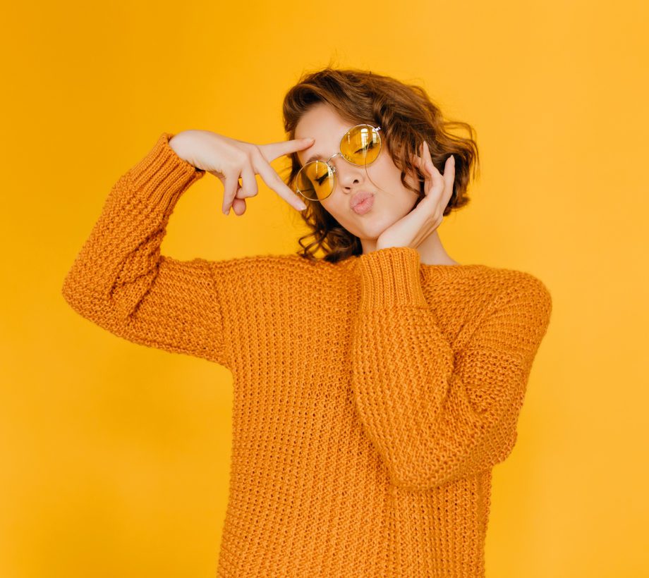 Dreamy girl in trendy sweater posing with kiss face expression and eyes closed. Lovely short-haired lady in glasses enjoying photoshoot on yellow background.